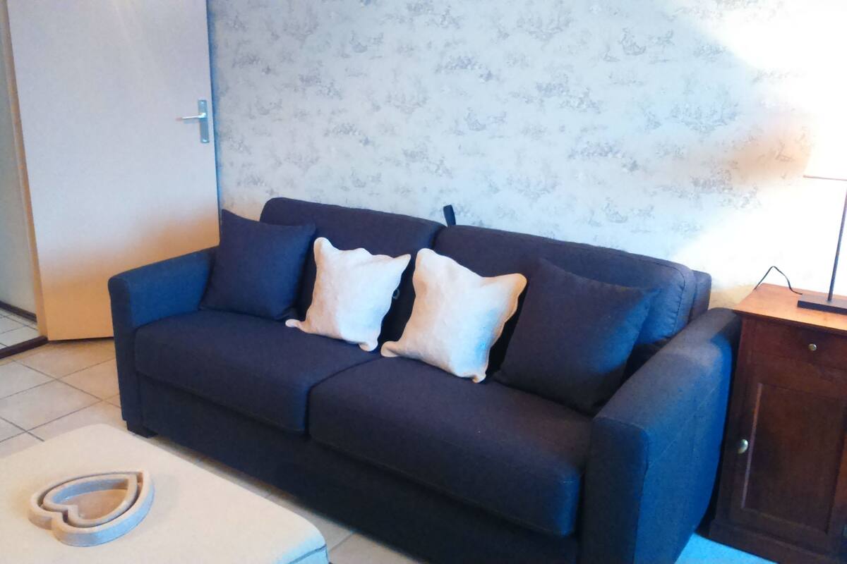 A living room with a dark blue sofa, two white cushions, a wooden side table, and a tiled floor.