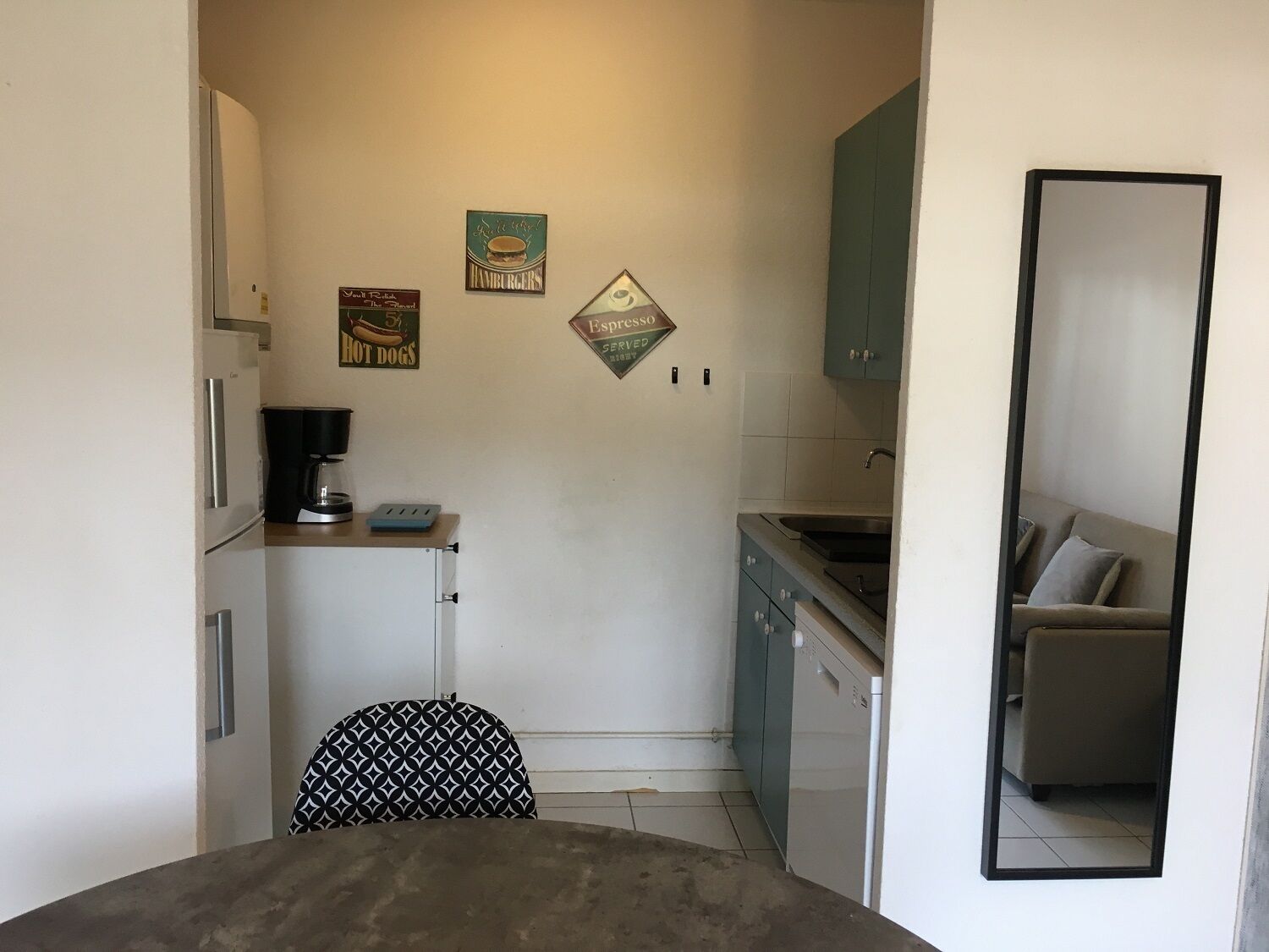 Een compacte keuken met een koelkast, koffiezetapparaat en een magnetron. Aan de muur decoraties, waaronder een bord met 'Hamburgers' en 'Espresso Served'. In de voorgrond staat een klein tafeltje en een stoel.