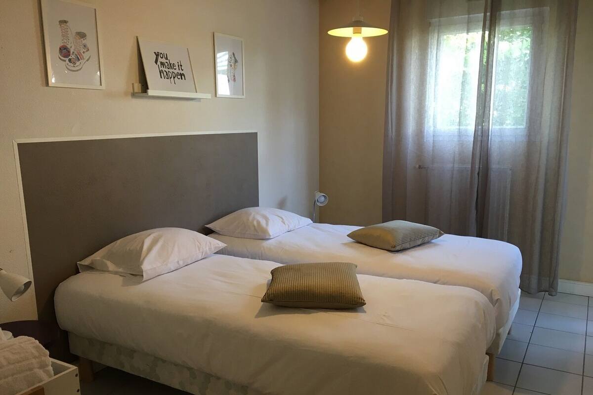 A hotel room with two beds, a headboard, a bedside table, and a window with curtains.