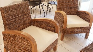 Two wicker chairs with cushions in a room with a tiled floor and a metal table with chairs in the background. - Tempologis - Meylan (Meylan)