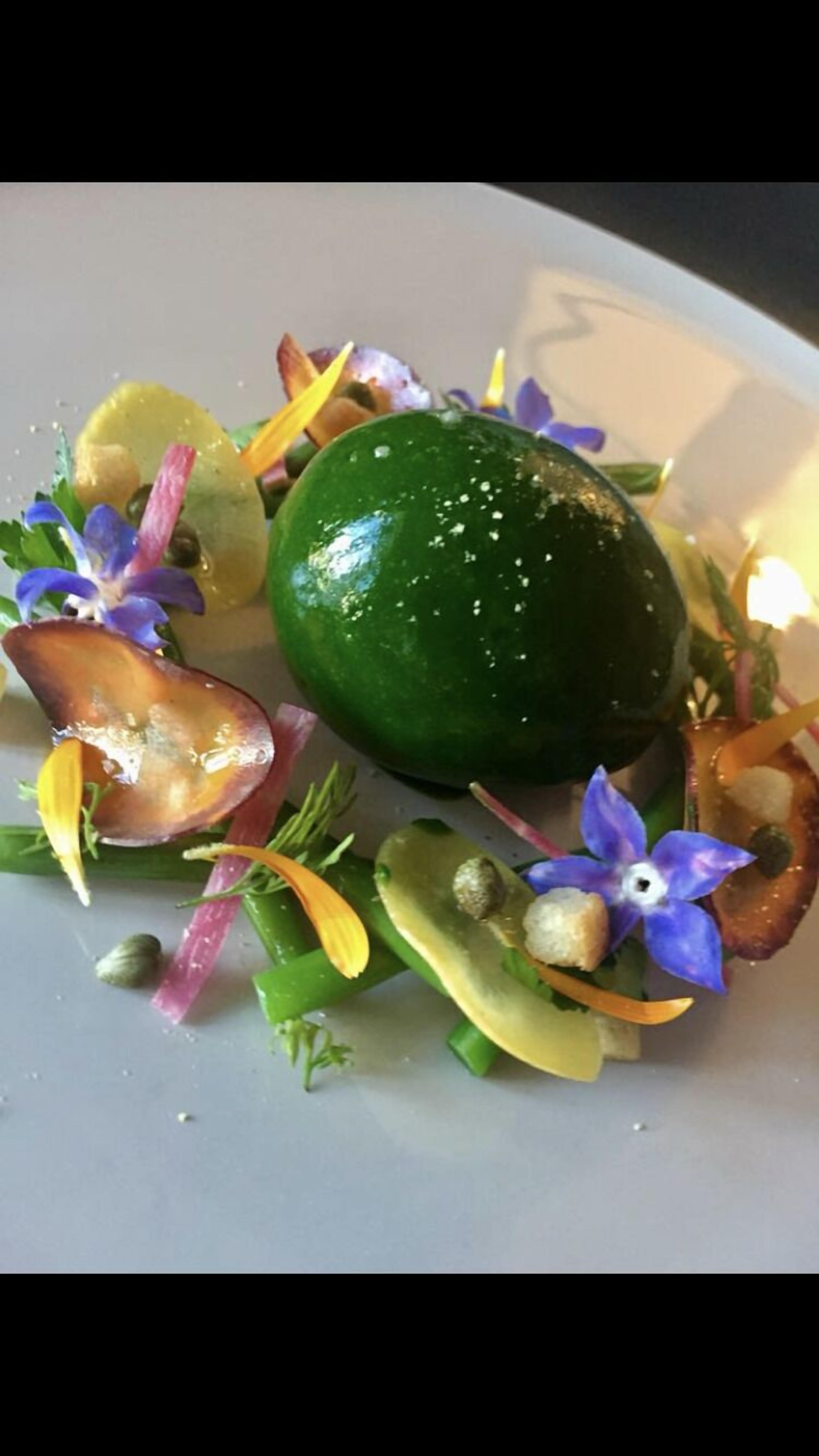 Un plato con un alimento esférico verde, rodeado de guarniciones y flores.