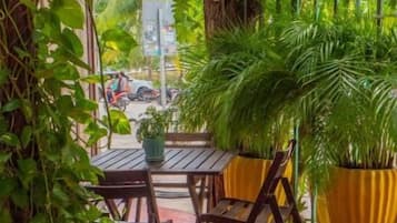 An outdoor seating area with tables and chairs, surrounded by lush greenery and a yellow awning.