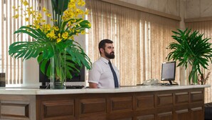 A reception desk with a man standing behind it, a large plant, and a computer monitor. - Pinhal Palace Hotel (Espirito Santo Do Pinhal)