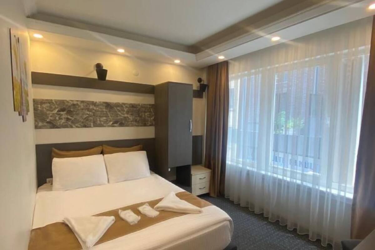 A hotel room with a large bed, a nightstand, a wall-mounted TV, and a window with sheer curtains.