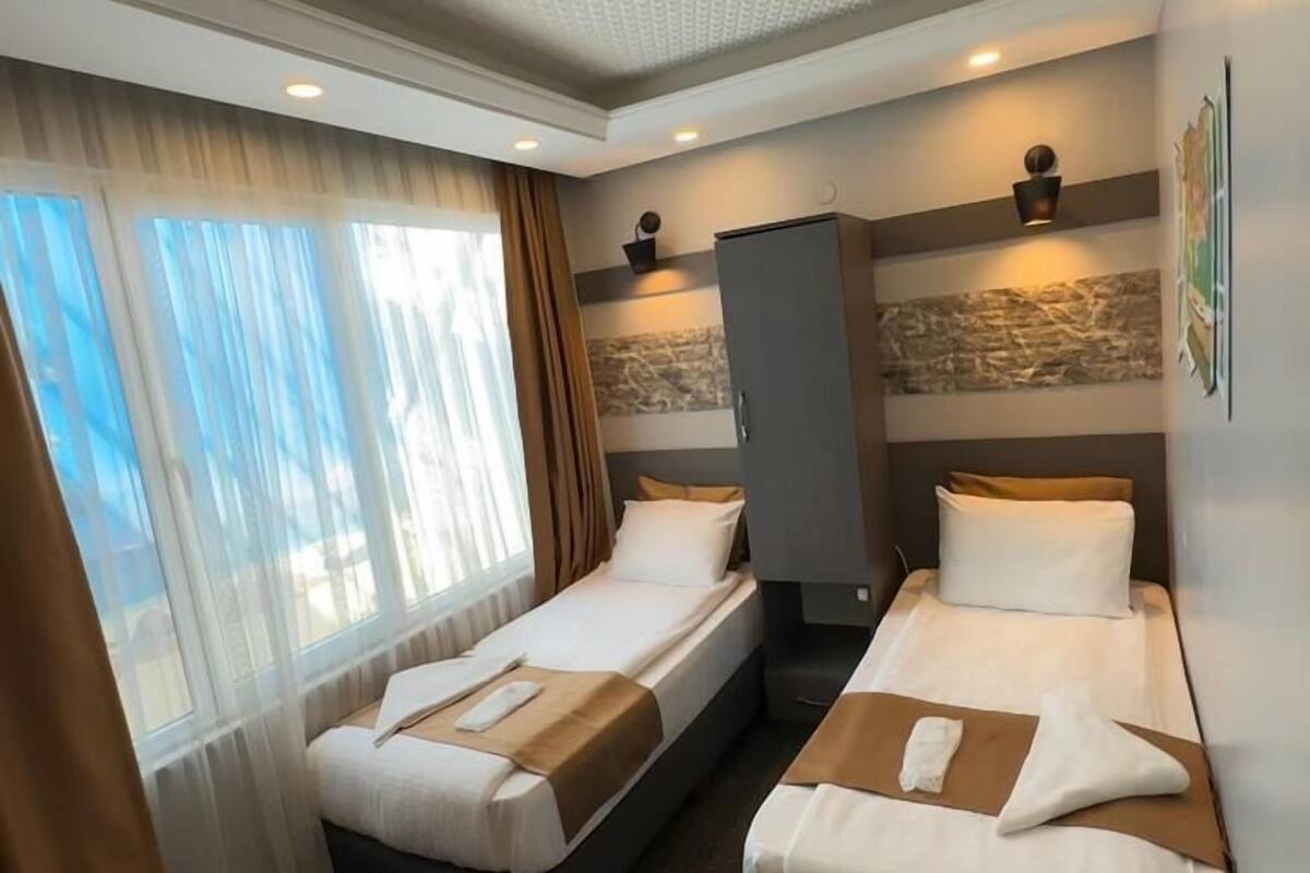 A hotel room with two single beds, a wardrobe, a large window with curtains, and a textured ceiling.