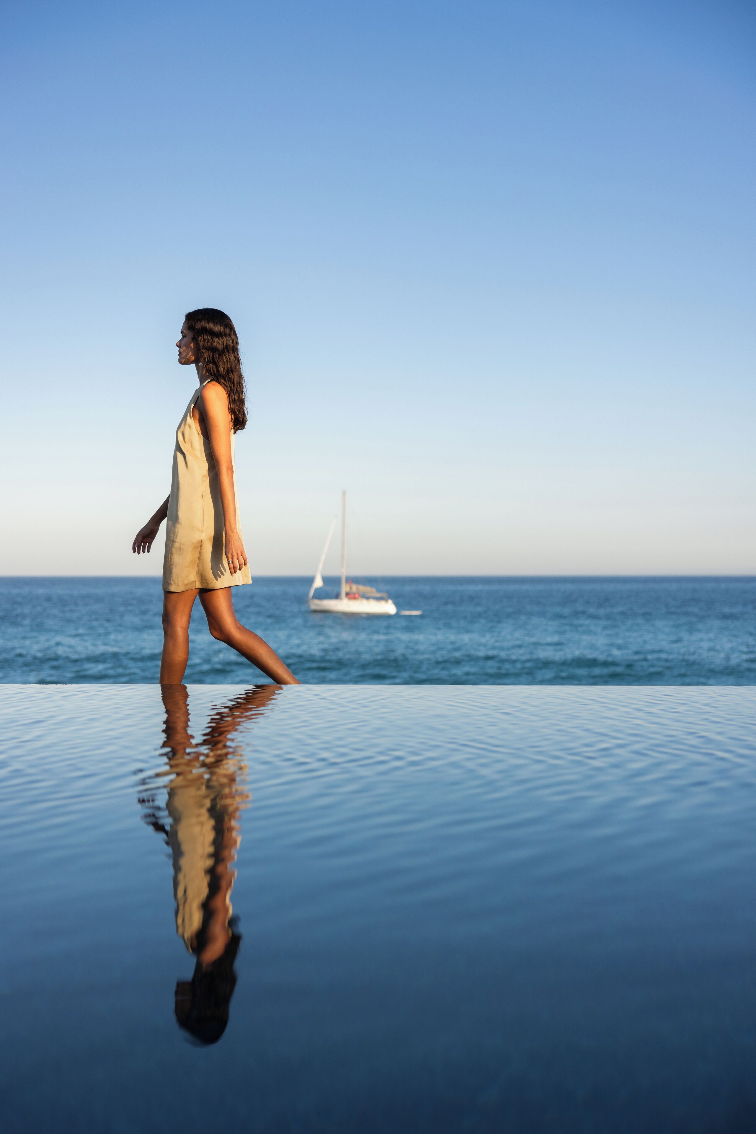 Una persona caminando sobre un borde de piscina reflectante, con un velero al fondo.