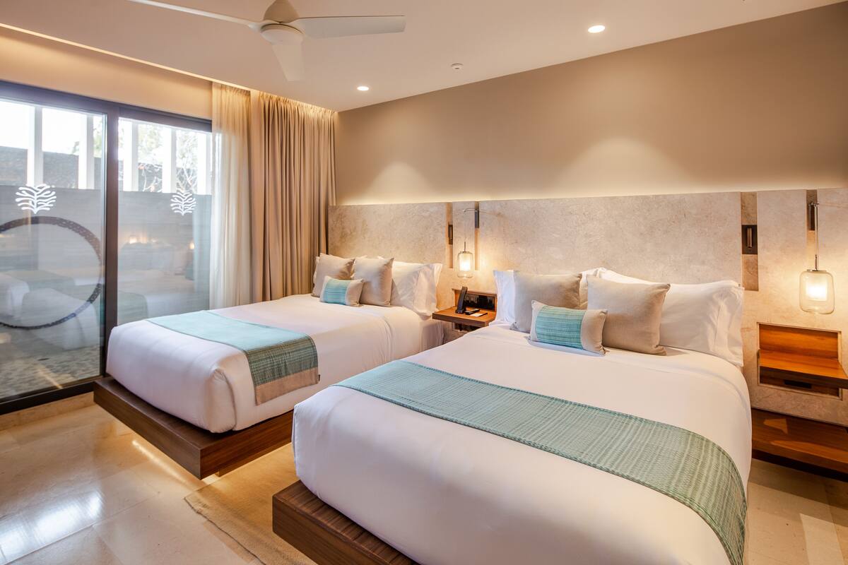 A modern hotel room with two beds, a large sliding glass door, and a ceiling fan.