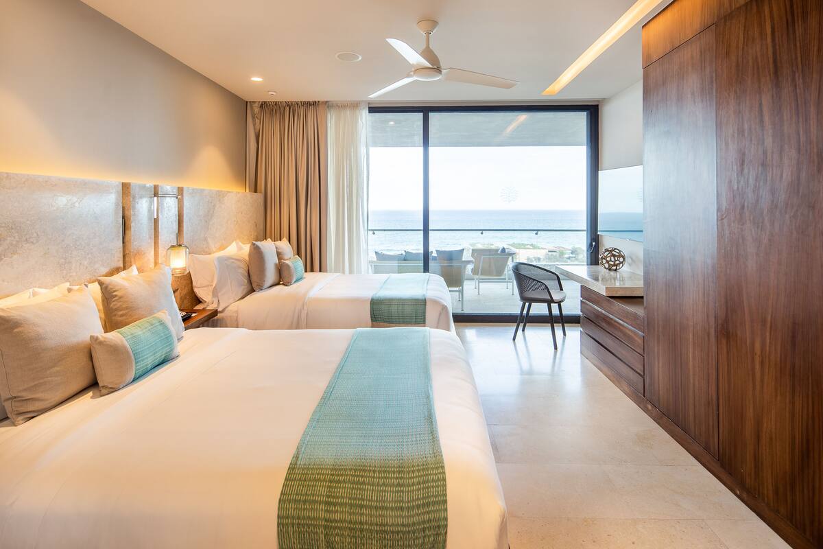 A hotel room with two beds, a balcony with a view, a desk, and a chair.