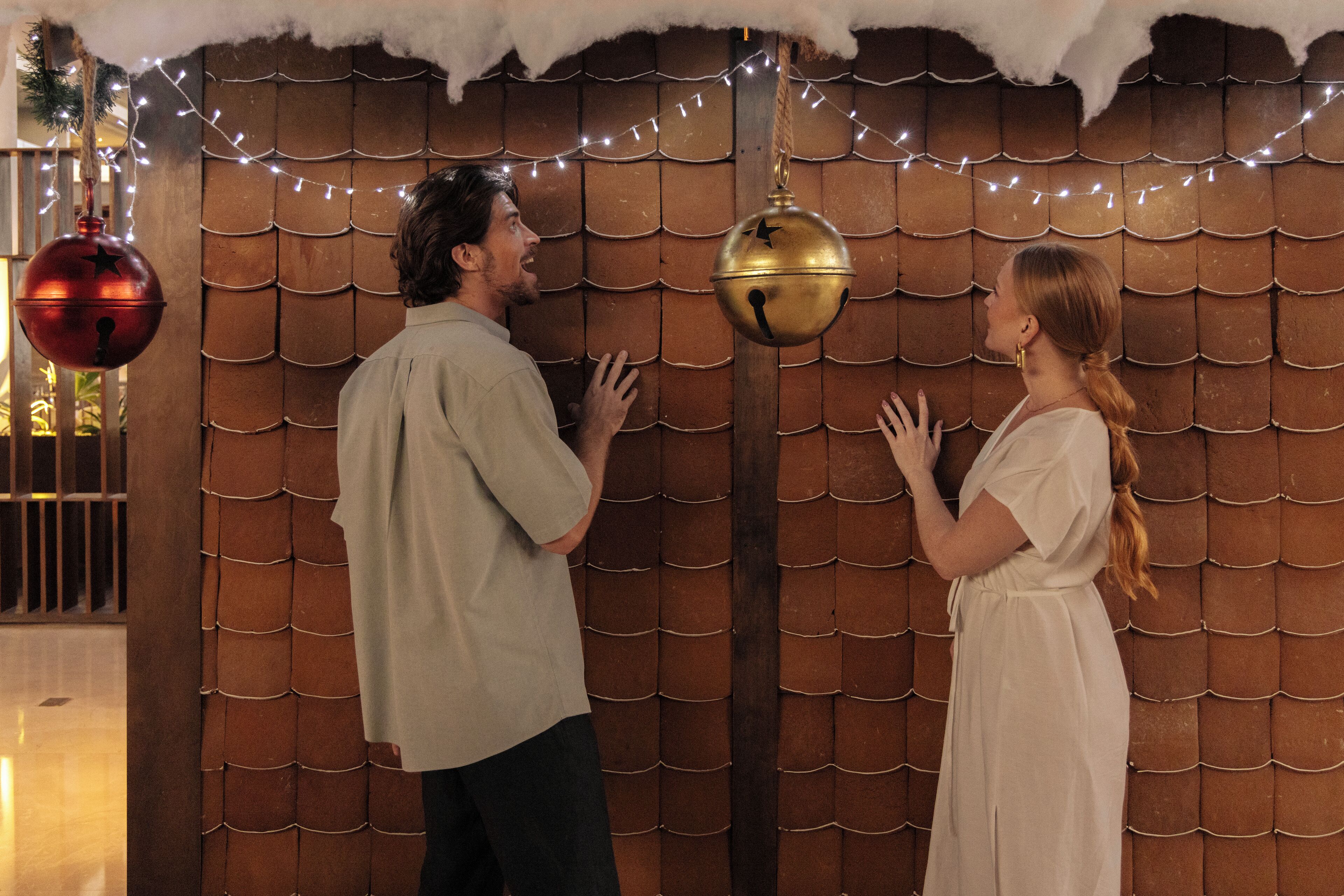 A man and a woman standing in front of a wooden wall with a scalloped pattern, decorated with string lights and hanging ornaments.