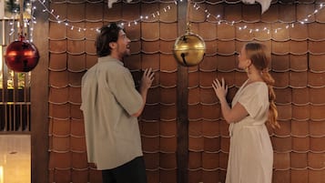 A man and a woman standing in front of a wooden wall with a scalloped pattern, decorated with string lights and hanging ornaments.
