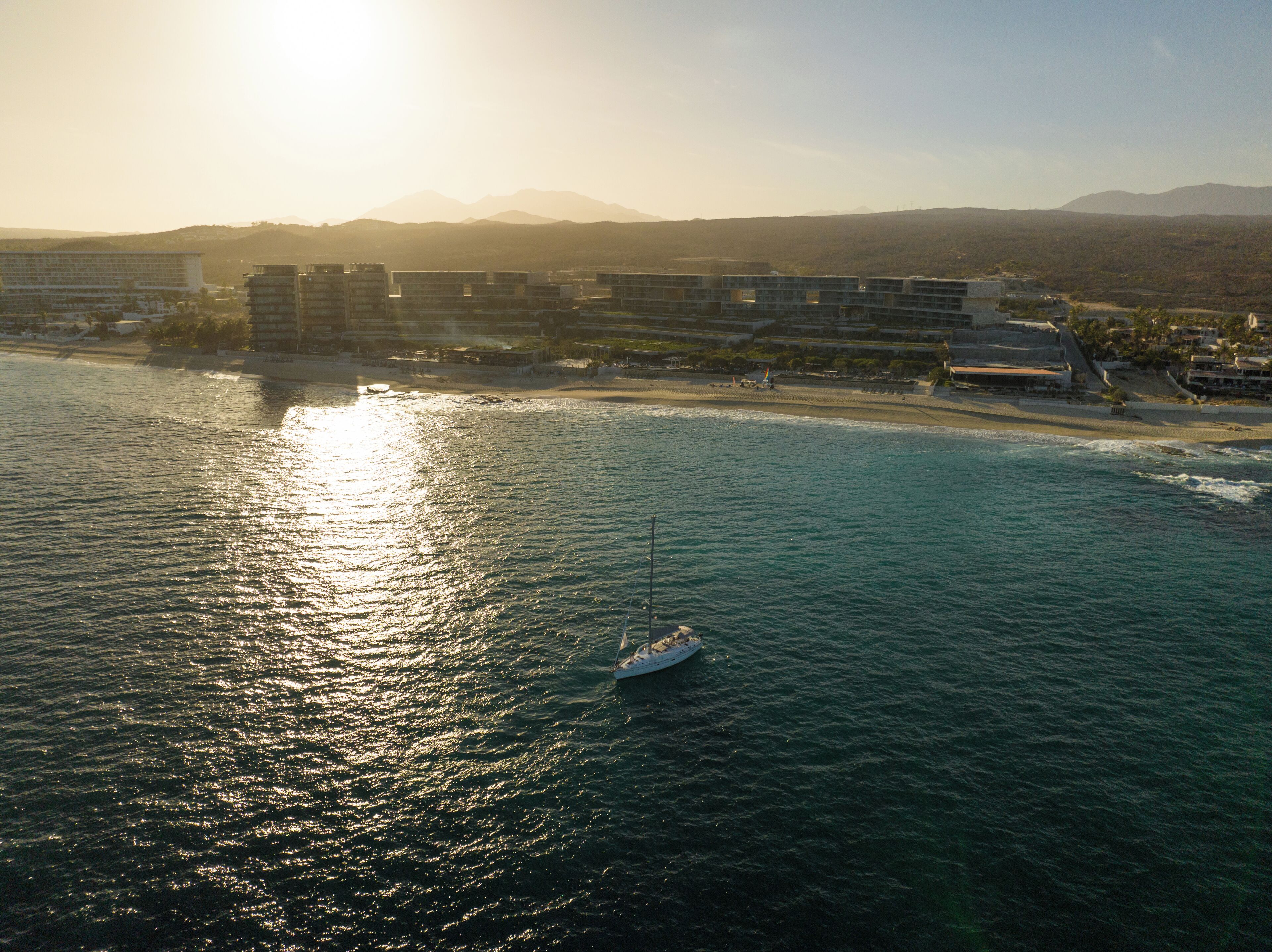 A coastal hotel with multiple buildings, a beach, and a sailboat in the water.