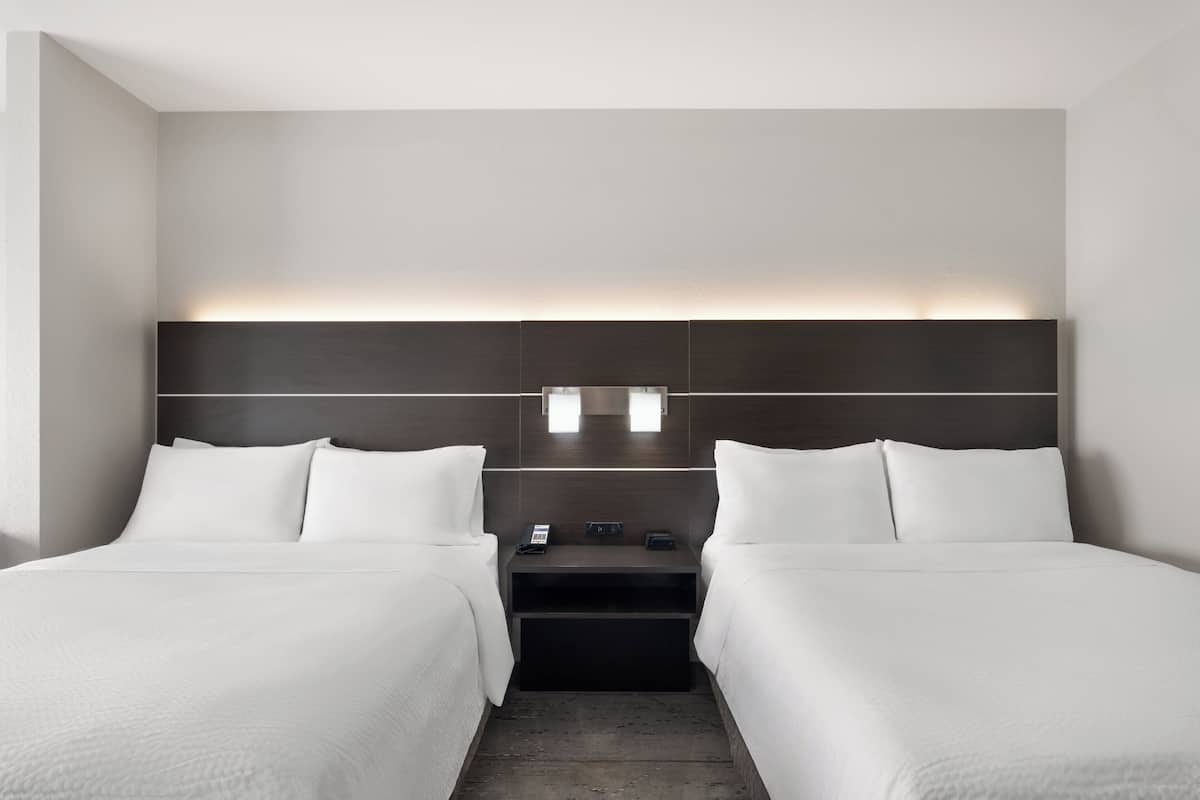 A hotel room with two single beds, a dark headboard, bedside tables with lamps, and a neutral color scheme.