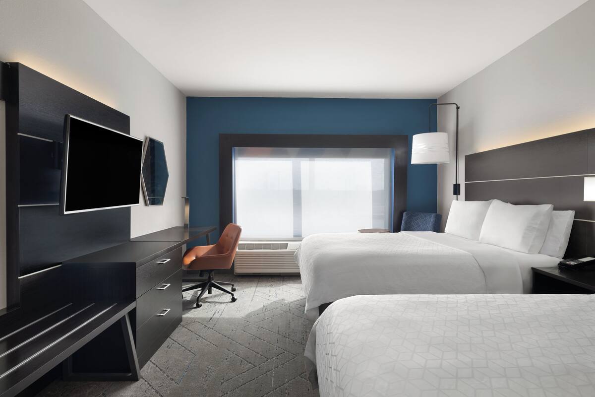 A modern hotel room with two beds, a desk, a chair, a TV, and a large window.
