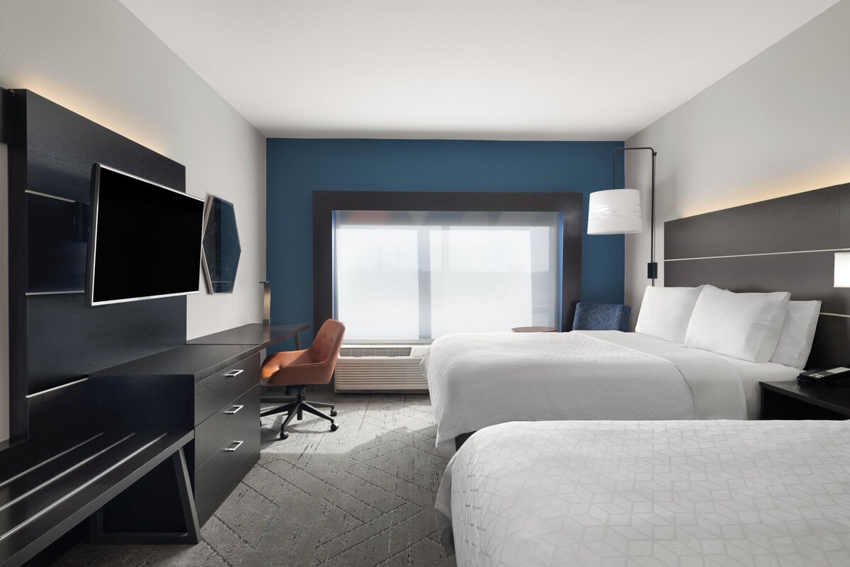 A modern hotel room with two beds, a desk, a chair, a TV, and a large window.