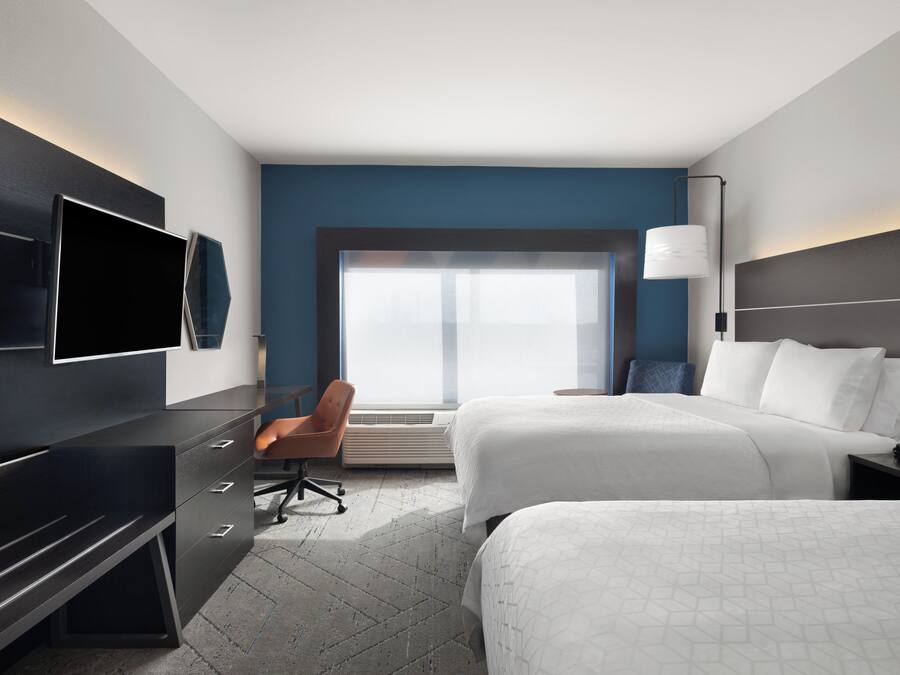 A modern hotel room with two beds, a desk, a chair, a TV, and a large window.