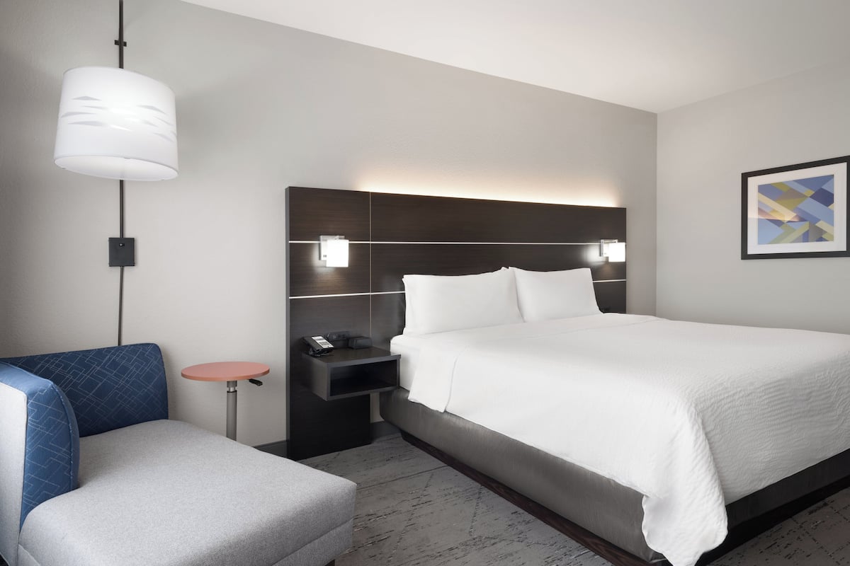 A modern hotel room with a large bed, a bedside table with a lamp, a small round table, and a blue armchair.