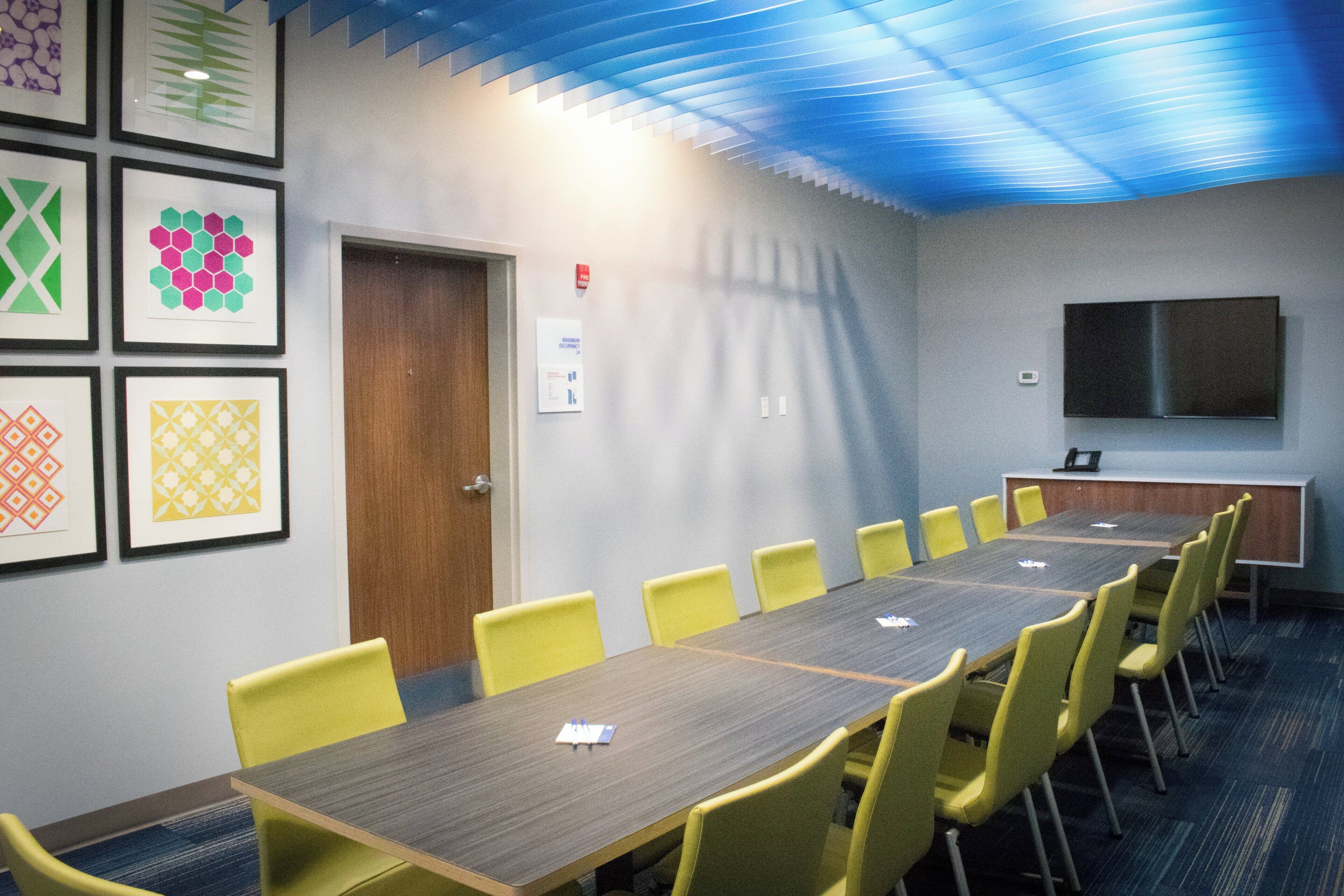 A conference room with a long table, yellow chairs, framed artwork on the wall, a door, and a mounted television.