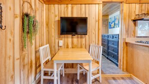 A wooden-paneled room with a TV mounted on the wall, a dining table with chairs, and a kitchen area with cabinets and a countertop. - 'Wapiti' Allenspark Cabin Near Rocky Mountains! (Allenspark)