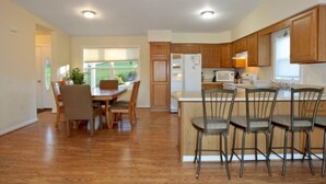 A modern kitchen with wooden cabinets, a central island with bar stools, a dining area with a table and chairs, and a view of the outdoors through large windows. - 5 o'clock Somewhere - Pool Table, Walking Distance to Portside Bar & Grill, Just Like New! (Moneta)