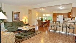 A living room with a green sofa, a coffee table, and a lamp. There is a dining area with a table and chairs. The kitchen has wooden cabinets and a refrigerator. - 5 o'clock Somewhere - Pool Table, Walking Distance to Portside Bar & Grill, Just Like New! (Moneta)