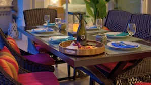 Una mesa de comedor dispuesta con platos, copas y una botella de vino, rodeada de sillas de mimbre y cojines.