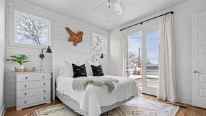 A bedroom with a large bed, white walls, a wooden dresser, a plant, and a view through the window.