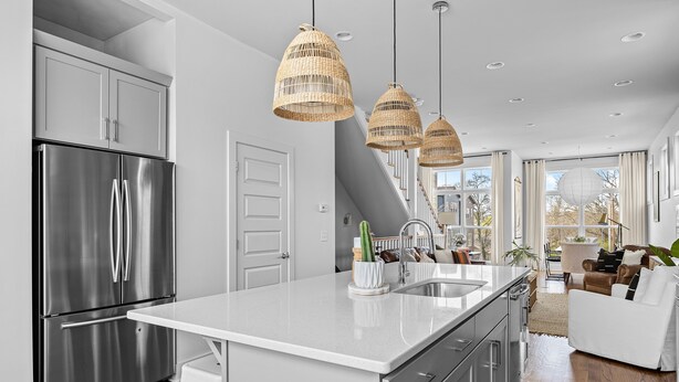 A modern kitchen with a stainless steel refrigerator, white cabinetry, and a large island with a sink. There are wicker pendant lights, a white countertop, and a view of the living area with a couch and a plant.