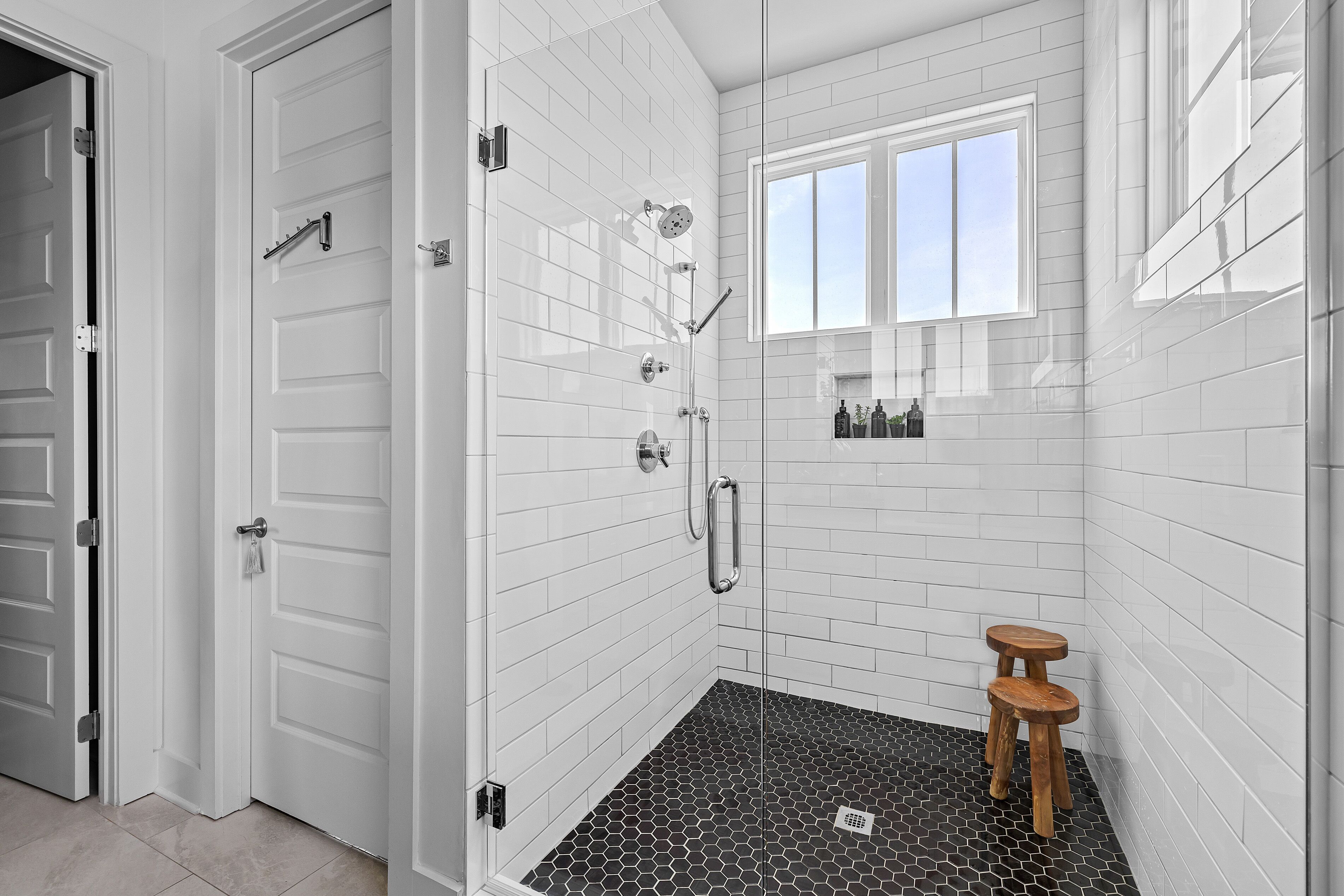 Une salle de bain moderne avec des murs recouverts de carreaux blancs, une douche en verre, un petit tabouret en bois et une fenêtre laissant entrer la lumière naturelle.