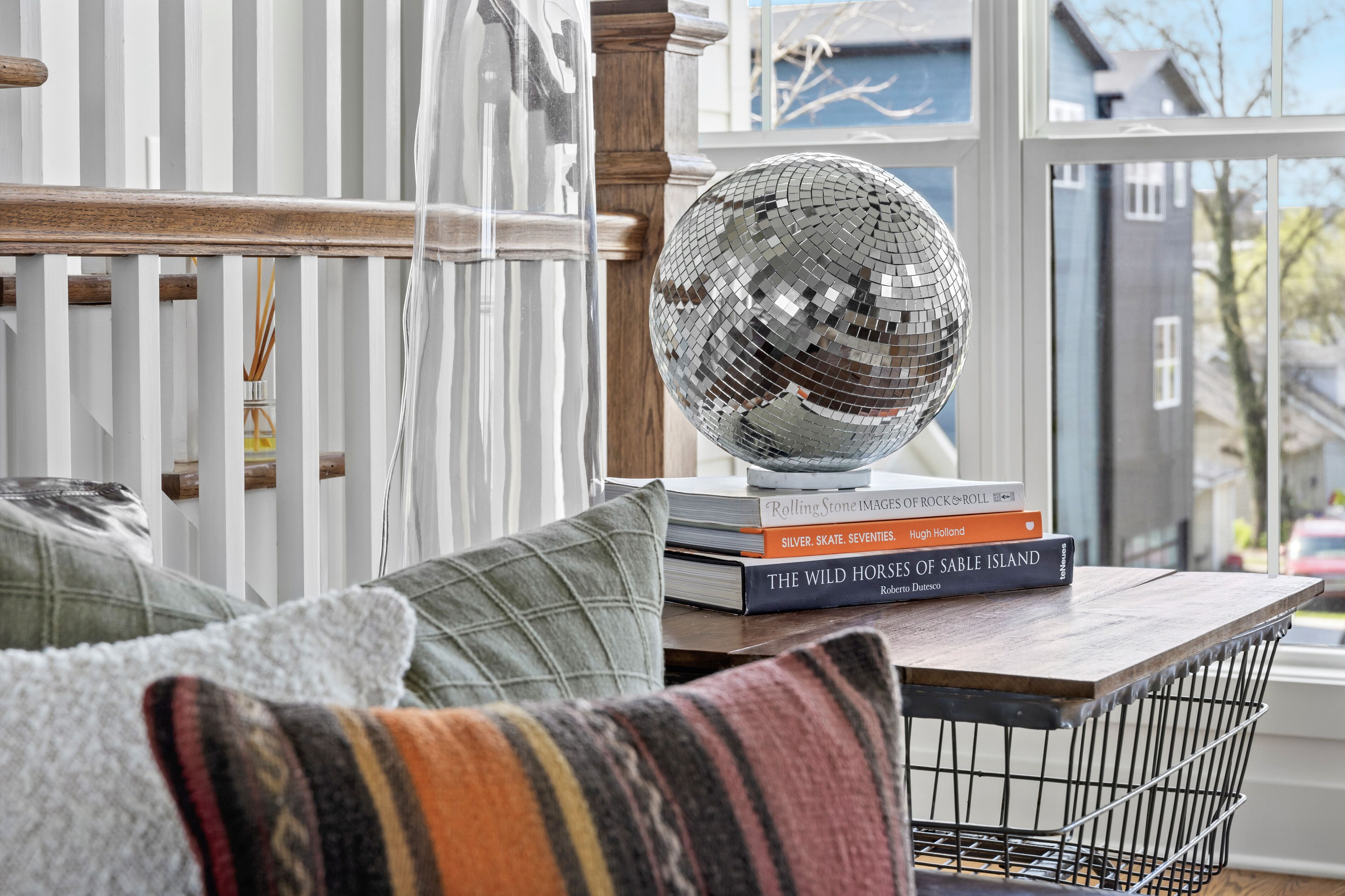 Ein Zimmer mit einem raumhohen Holzrahmenfenster, einem Drahtkorb und einem Stapel Bücher auf einem Holztisch.