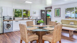 Une cuisine moderne avec des meubles blancs, un lave-linge encastré et un coin repas agrémenté de chaises en osier et d’une table en verre.