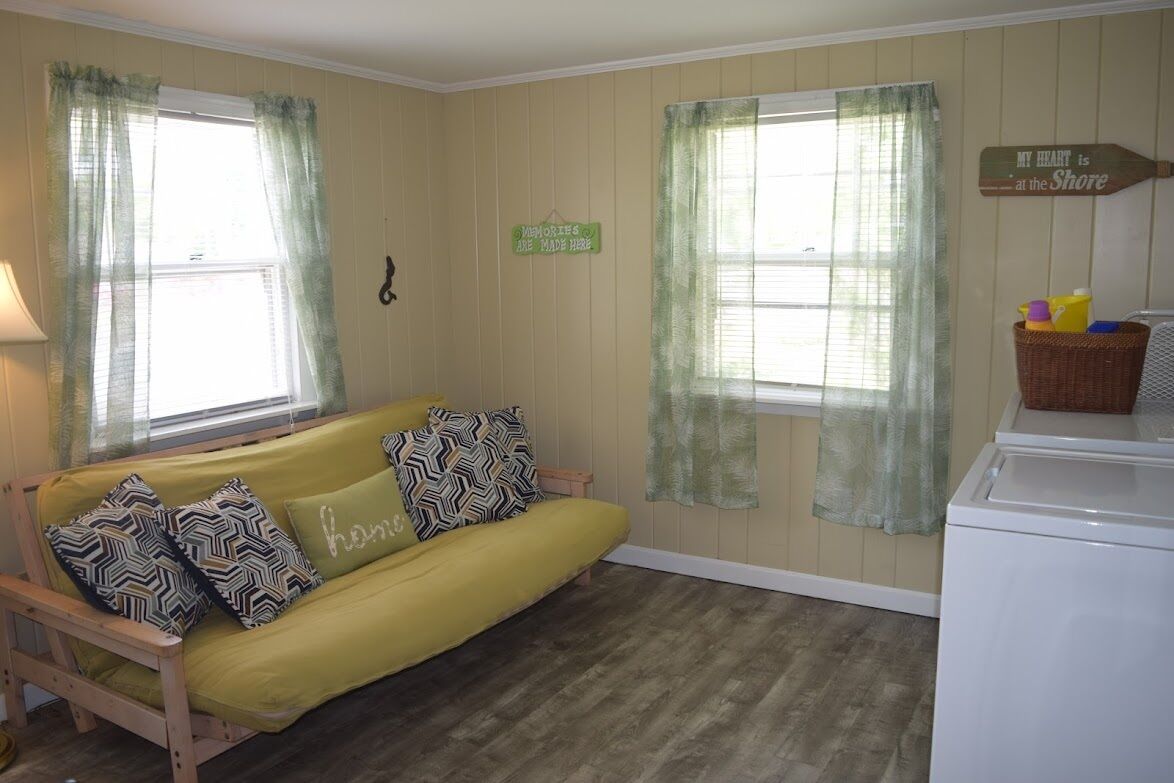 Um quarto com sofá amarelo, duas janelas com cortinas leves, uma placa na parede, uma máquina de lavar e uma pequena mesa com um cesto.