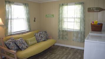 Um quarto com sofá amarelo, duas janelas com cortinas leves, uma placa na parede, uma máquina de lavar e uma pequena mesa com um cesto.