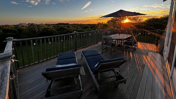 A wooden deck with lounge chairs and a table, overlooking a sunset.