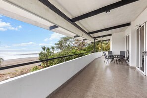Un balcón con vistas a la playa, una mesa con sillas y una puerta de cristal que da a un espacio interior.