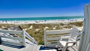 Blick auf einen Strand von einer Holzdielen-Terrasse mit weißen Geländern und Stühlen, mit Blick auf einen Sandstrand, auf dem sich Menschen und Sonnenschirme befinden.