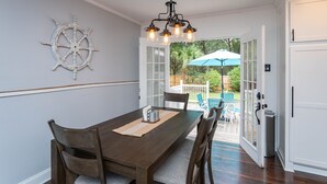 Un comedor con una mesa y sillas de madera, un adorno de timón y vista a un patio trasero con sombrilla.