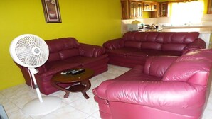 A living room with a sectional sofa, a standing fan, and a coffee table. - BPO/Business Travellers/ Large Group Accommodation  (Spanish Town)