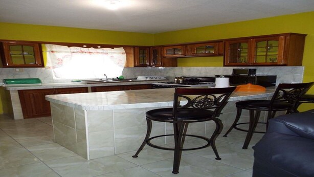A kitchen with a central island, two bar stools, wooden cabinets, and a window with curtains. - BPO/Business Travellers/ Large Group Accommodation  (Spanish Town)