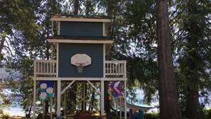 Une cabane à jouer avec un panier de basket, entourée d’arbres et bordée par un plan d’eau.