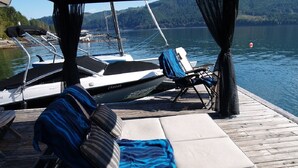 Une terrasse dotée d’un canapé, de chaises longues et d’un espace couvert offrant une vue sur les bateaux et un lac.