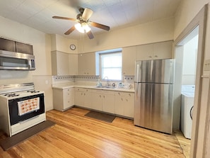 A kitchen with wooden flooring, a stove, a microwave, a refrigerator, and a ceiling fan.