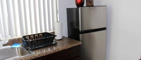 A kitchen with a stainless steel refrigerator, a granite countertop, a sink, and a window with blinds.