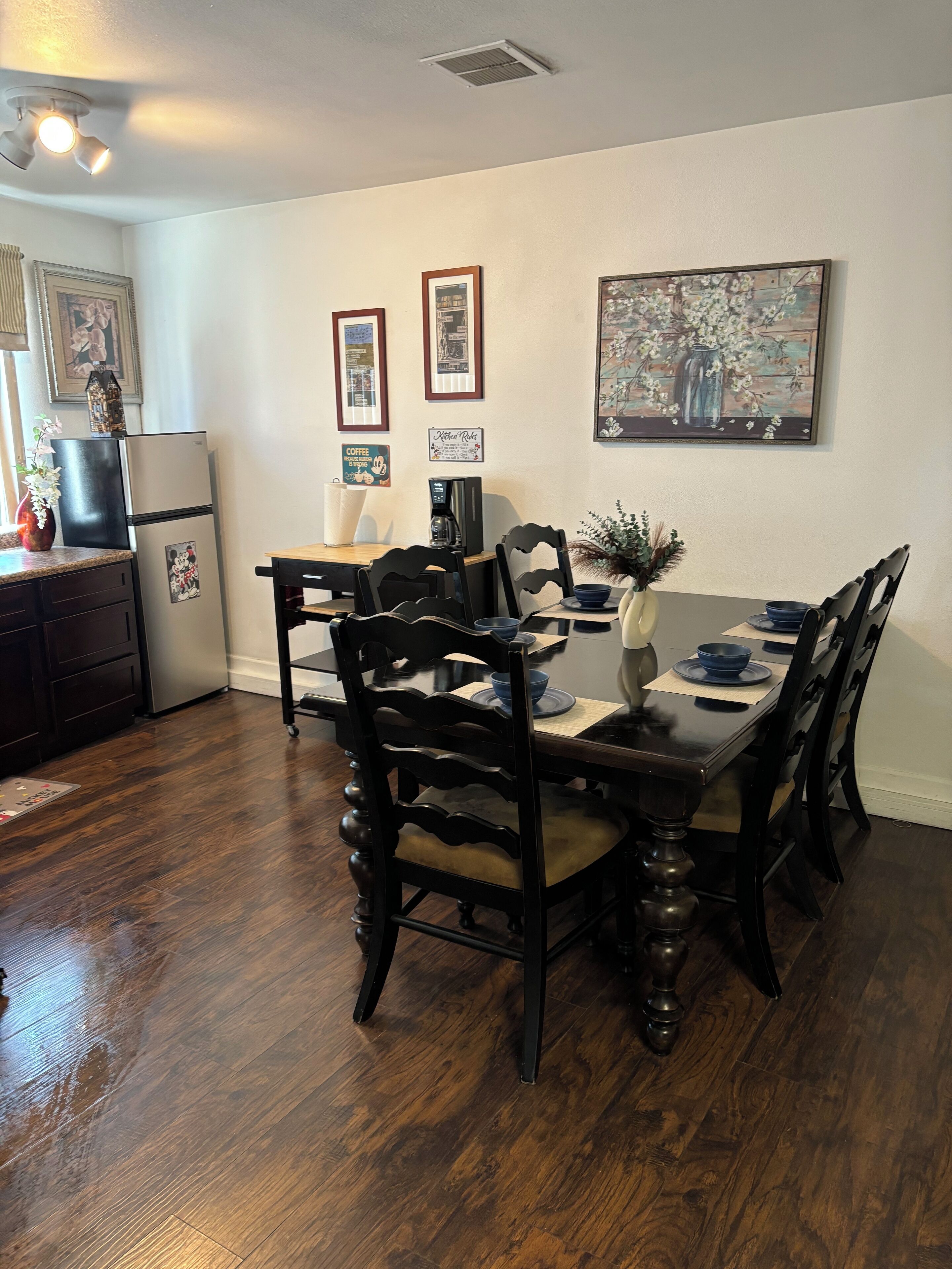 A dining area with a wooden table and chairs, a refrigerator, and framed pictures on the wall.