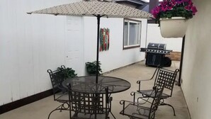 A patio with a round glass table and four chairs, an umbrella, and a grill in the background.