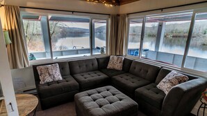 Una sala de estar acogedora con un sofá seccional y reposapiés, ventanas grandes con cortinas y vistas a un lago.