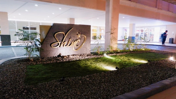 Un letrero con la palabra 'Shine' iluminada en letras doradas, rodeado por un área ajardinada con piedras y césped, y una persona de pie al fondo.