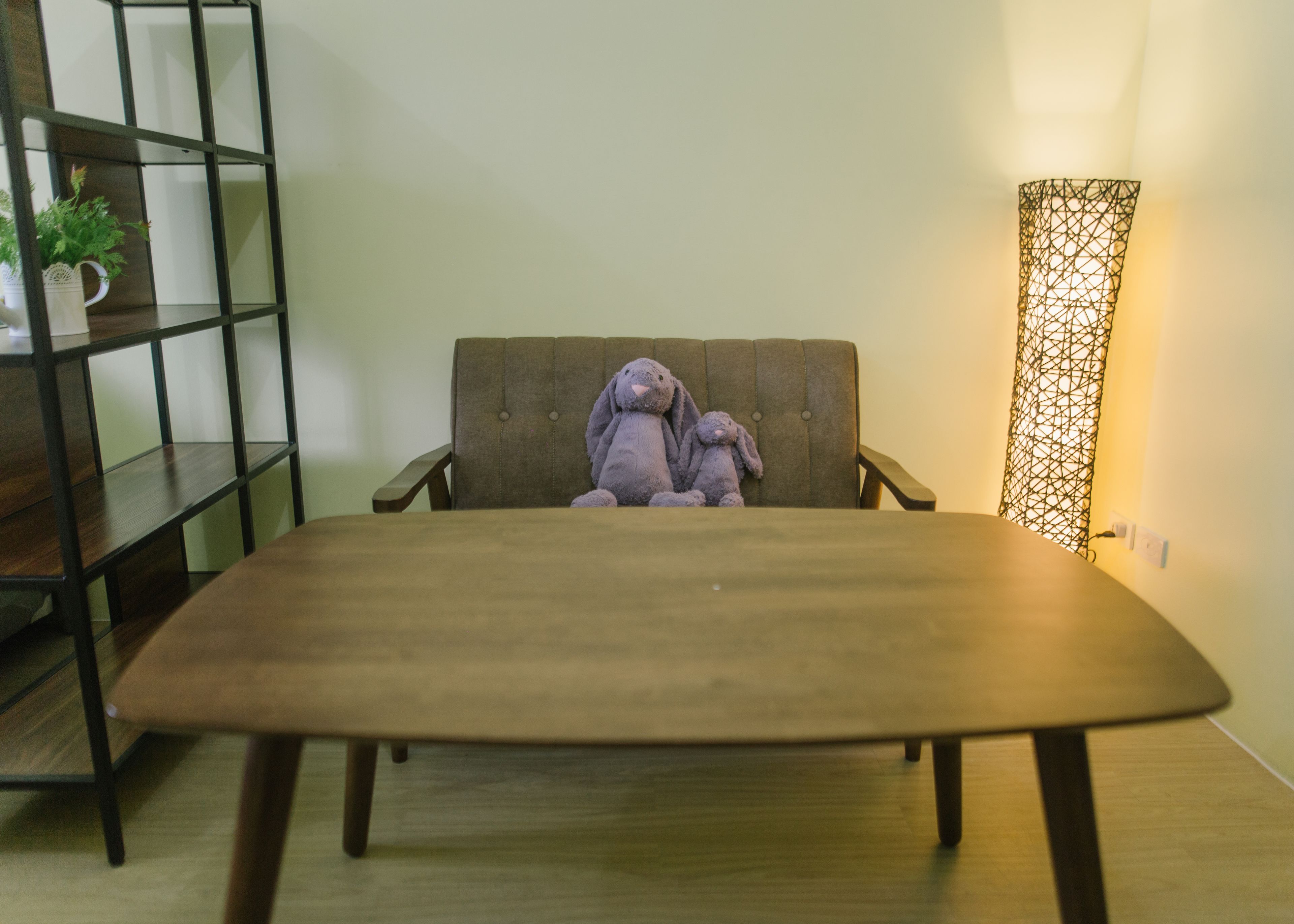 A room with a wooden table, a grey sofa, a black metal shelf with a plant, and a wall-mounted light fixture.