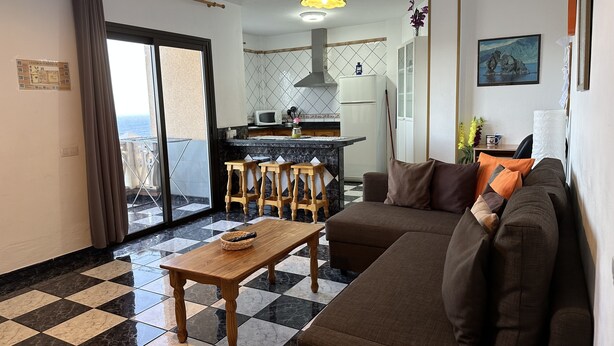 Sala de estar com sofá, mesa de madeira e área de cozinha com um banquinho.