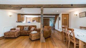A cozy living room with a brown leather sofa set, wooden dining table, and chairs. A wooden beam supports the ceiling, and there is a kitchen area visible in the background.