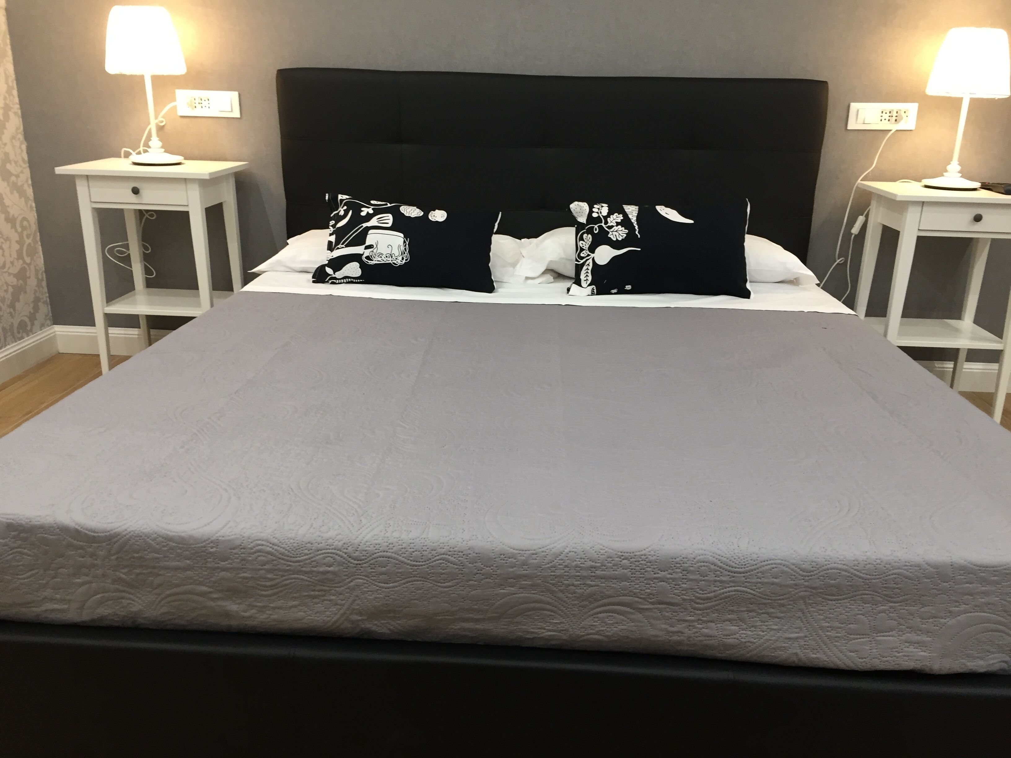 Una cama bien hecha con un colchón blanco y dos almohadas negras con diseños blancos, flanqueada por dos mesitas de noche con lámparas, en una habitación con paredes grises y suelo de madera.